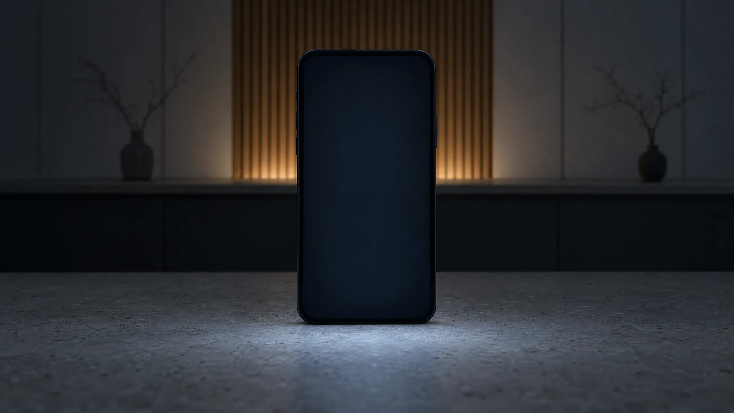 A black phone standing on the pale stone worktop of a display kitchen at night, its screen lighting the stone; behind it, out of focus, a slatted oak panel lit from below and a vase with a bare branch on either side