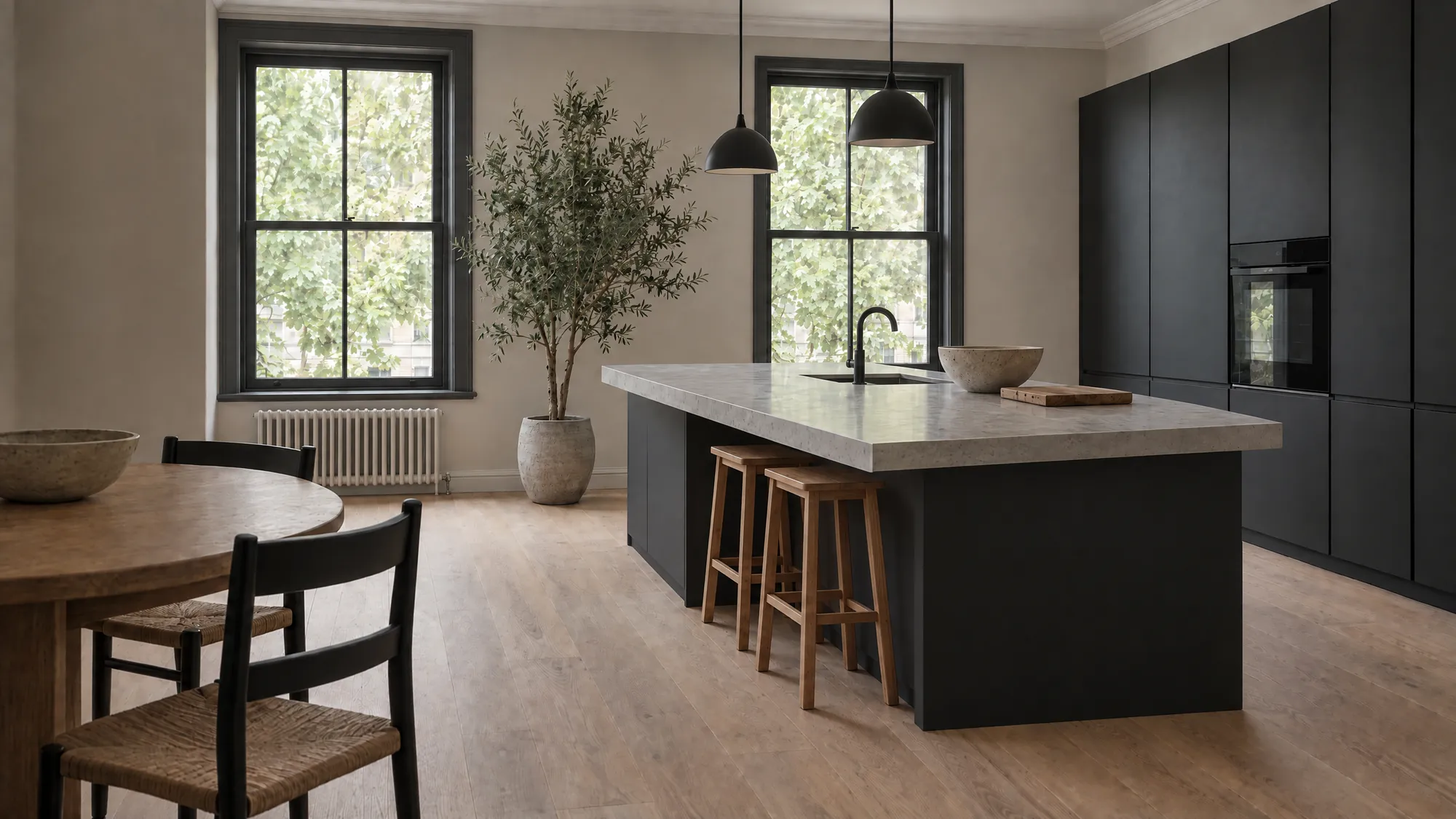 A kitchen in a converted Victorian flat: a long matte black island with a thick pale grey stone top and two oak stools under two black pendant lights, black tall units with a built-in oven, two tall black sash windows onto plane trees, an olive tree, and a round oak table in the foreground